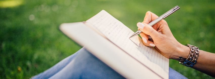 Osoba pisząca długopisem w notatniku na kolanach, siedząca na trawie w parku. W tle rozmyta zieleń, a na ręce osoby widać bransoletki.